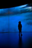 A silhouette of a person stands in front of a large screen displaying shades of blue and textured patterns, creating a mysterious and immersive atmosphere.