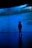 A silhouette of a person stands in front of a large screen displaying shades of blue and textured patterns, creating a mysterious and immersive atmosphere.