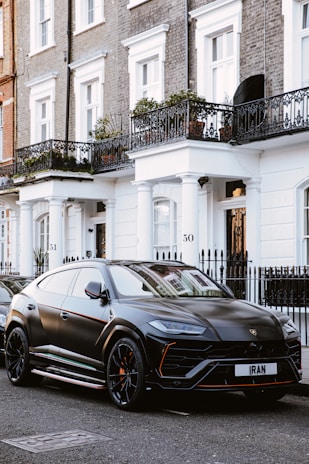 A sleek, black luxury SUV is parked on a street with elegant, brick townhouses in the background. The vehicle's design is modern and sporty, with tinted windows and distinctive branding. The backdrop features white-trimmed windows and ornate wrought-iron balconies adorned with potted plants.
