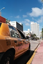 A yellow taxi is parked on the side of a street with modern buildings lining the background. One of the buildings is tall and cylindrical, while others are rectangular and varied in height. There are palm trees along the street, and the sky is blue with a few clouds.