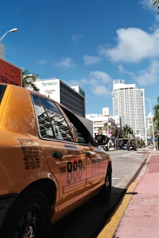 A yellow taxi is parked on the side of a street with modern buildings lining the background. One of the buildings is tall and cylindrical, while others are rectangular and varied in height. There are palm trees along the street, and the sky is blue with a few clouds.