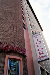 An upward view of a tall building with a textured facade and vertical pink accents. A prominent 'Liquor' sign is displayed, with a neon sign visible in the window below. The building has numerous narrow windows aligned in a column.