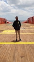 A person stands on the deck of a large ship or maritime platform, with the ocean visible in the background and a cloudy sky above. The deck is wooden with yellow markings and red safety structures on either side.