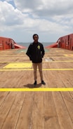 A person stands on the deck of a large ship or maritime platform, with the ocean visible in the background and a cloudy sky above. The deck is wooden with yellow markings and red safety structures on either side.