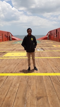 A person stands on the deck of a large ship or maritime platform, with the ocean visible in the background and a cloudy sky above. The deck is wooden with yellow markings and red safety structures on either side.