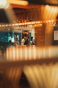A bartender stands behind a counter, surrounded by inverted wine glasses hanging above. The setting is dimly lit, with a warm ambiance created by multiple small lights. Reflections and blurred elements in the foreground add depth to the scene.