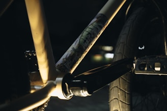 Close-up view of a bicycle frame with a focus on the crankset area. The frame has artistic graffiti-like designs and is lit dramatically with shadows. The background is blurred, emphasizing the bike components.