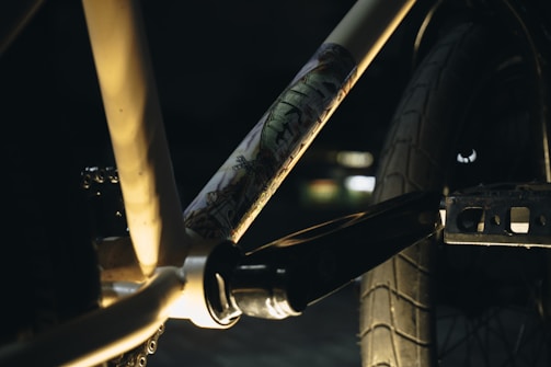Close-up view of a bicycle frame with a focus on the crankset area. The frame has artistic graffiti-like designs and is lit dramatically with shadows. The background is blurred, emphasizing the bike components.