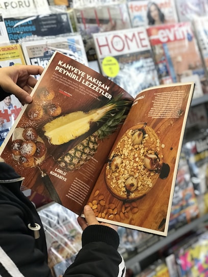 A person is holding an open magazine featuring a section on food. The left page displays images of a pineapple and toasted bread rolls on a brown surface. The right page shows a large baked dish topped with oats and nuts. In the background, there is a display of various magazines.