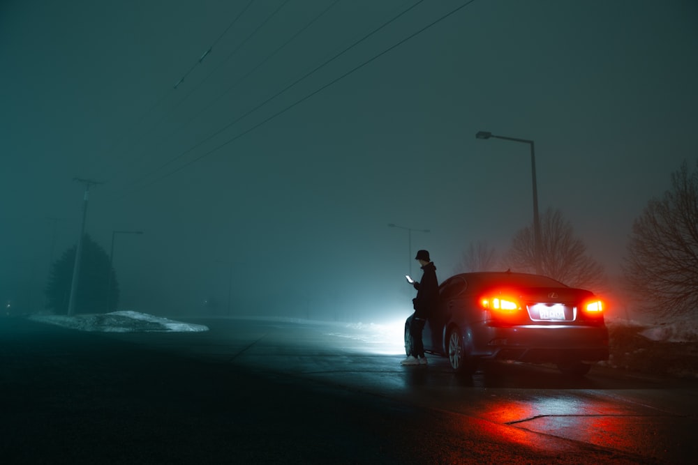 man in black jacket and black pants standing beside red car during night time