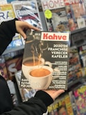 A person holding a magazine titled 'Kahve' shows a close-up of a coffee machine pouring espresso into a white cup. It features articles about franchising coffee shops and various coffee-related topics. In the background, there is a display of various other magazines on shelves.