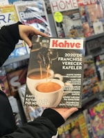 A person holding a magazine titled 'Kahve' shows a close-up of a coffee machine pouring espresso into a white cup. It features articles about franchising coffee shops and various coffee-related topics. In the background, there is a display of various other magazines on shelves.