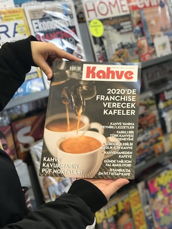 A person holding a magazine titled 'Kahve' shows a close-up of a coffee machine pouring espresso into a white cup. It features articles about franchising coffee shops and various coffee-related topics. In the background, there is a display of various other magazines on shelves.