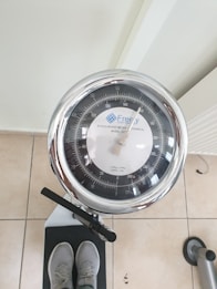 A mechanical scale with a round dial, displaying weight measurements in kilograms and pounds. The scale is positioned on a tiled floor and a pair of feet wearing gray shoes are visible on the scale's platform. The dial features a chrome rim and a needle indicating the weight measurement.
