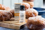 A bottle of grapefruit flavoring is placed on a wooden board among sugared doughnuts. The board has a slightly rustic appearance, and the doughnuts are drizzled with a glaze. One doughnut is prominently placed behind the flavoring bottle.