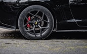 Close-up of a black car's tire and alloy wheel, with red brake calipers visible. The vehicle is parked on a textured asphalt surface with some scattered leaves.