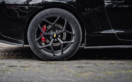 Close-up of a black car's tire and alloy wheel, with red brake calipers visible. The vehicle is parked on a textured asphalt surface with some scattered leaves.