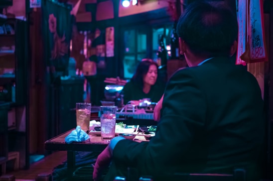 A dimly lit restaurant or bar setting with a person in a suit seated at the table, facing a woman who is slightly out of focus. The table has drinks and plates with food, and the ambiance is characterized by low blue and pink lighting.