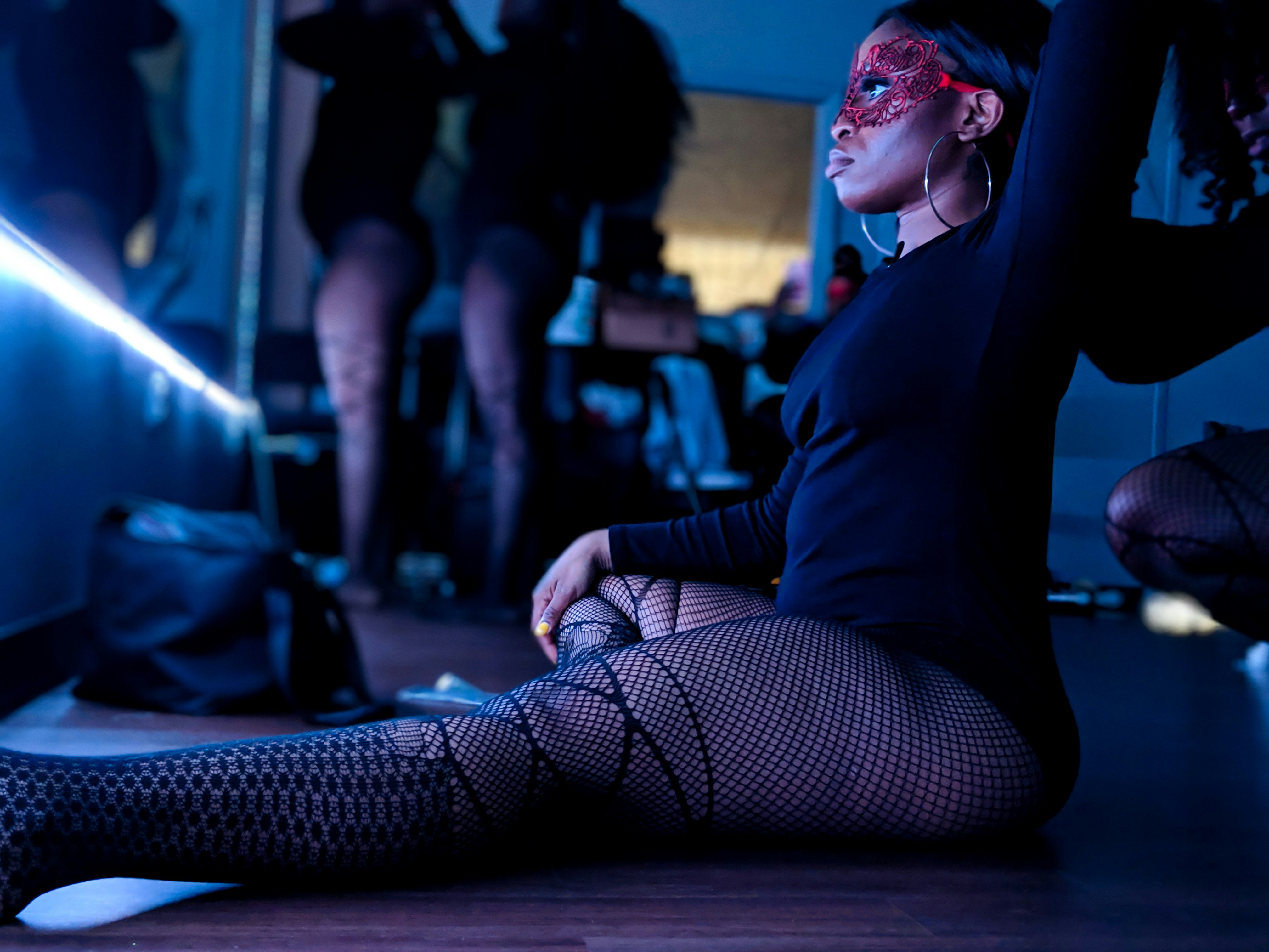 A dancer in fishnet stockings sits in front of a lighted mirror while a coworker styles her hair. 
