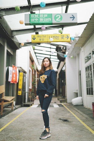 A young woman stands in an urban passageway, wearing a navy blue sweatshirt with a lion cub graphic and ripped jeans. Overhead signs are written in Korean, with decorative paper lanterns hanging from the ceiling structure. The passageway has a mix of modern and casual elements, creating a relaxed atmosphere.