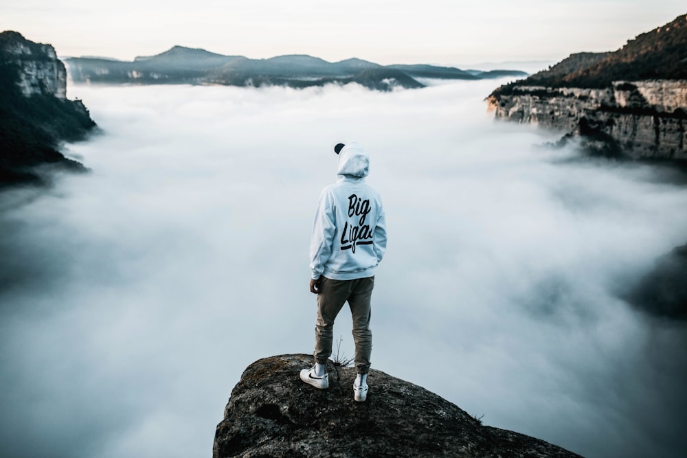 man in white hoodie standing on rock near body of water during daytime