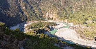 A meandering river flows through a lush, forested valley. The river's vibrant blue-green waters contrast with the rocky, beige riverbanks. Steep, densely vegetated hills rise on both sides, forming a natural enclosure around the waterway. Sparse patches of gravel and small shrubs are scattered throughout the landscape.