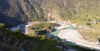A meandering river flows through a lush, forested valley. The river's vibrant blue-green waters contrast with the rocky, beige riverbanks. Steep, densely vegetated hills rise on both sides, forming a natural enclosure around the waterway. Sparse patches of gravel and small shrubs are scattered throughout the landscape.