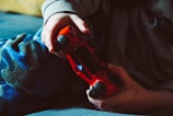 A person is holding a red gaming controller while sitting on a couch, wearing comfortable clothing. The scene is dimly lit, giving a relaxed atmosphere.