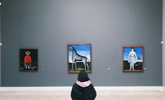 A person is seated in an art gallery facing three paintings on a neutral-colored wall. The paintings depict unique, surreal scenes: a figure in a red outfit and hat, a horse-like creature with a long neck against a landscape, and a woman in a dress holding a small creature.
