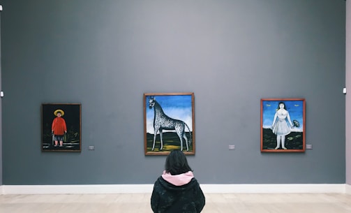 A person is seated in an art gallery facing three paintings on a neutral-colored wall. The paintings depict unique, surreal scenes: a figure in a red outfit and hat, a horse-like creature with a long neck against a landscape, and a woman in a dress holding a small creature.