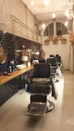 A stylish, modern barbershop interior with black leather chairs and a wooden counter. The walls feature black tiles and exposed pipes, with hanging lights providing a warm ambiance. Shelving holds various grooming products, and a large, arched window is visible at the back.