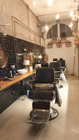 A stylish, modern barbershop interior with black leather chairs and a wooden counter. The walls feature black tiles and exposed pipes, with hanging lights providing a warm ambiance. Shelving holds various grooming products, and a large, arched window is visible at the back.