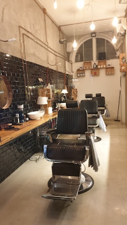 A stylish, modern barbershop interior with black leather chairs and a wooden counter. The walls feature black tiles and exposed pipes, with hanging lights providing a warm ambiance. Shelving holds various grooming products, and a large, arched window is visible at the back.