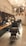 A stylish, modern barbershop interior with black leather chairs and a wooden counter. The walls feature black tiles and exposed pipes, with hanging lights providing a warm ambiance. Shelving holds various grooming products, and a large, arched window is visible at the back.