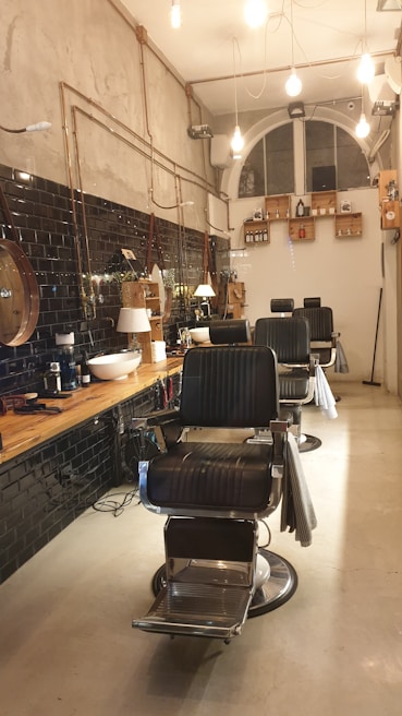 A stylish, modern barbershop interior with black leather chairs and a wooden counter. The walls feature black tiles and exposed pipes, with hanging lights providing a warm ambiance. Shelving holds various grooming products, and a large, arched window is visible at the back.