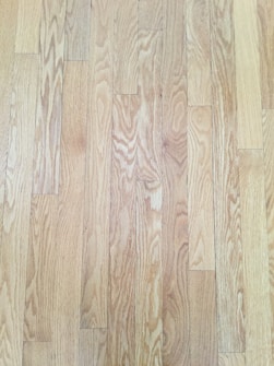 A close-up view of wooden floorboards, showing a natural wood grain pattern with varying shades of light brown and beige. The floorboards are aligned in parallel rows, exhibiting a smooth and polished surface.