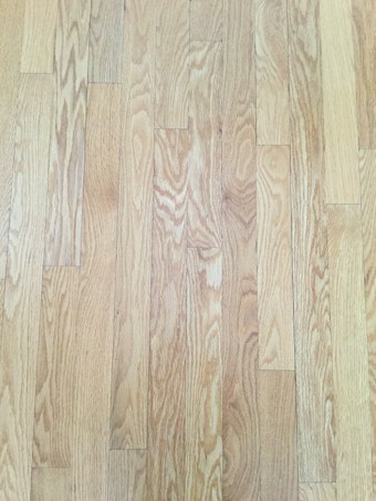 A close-up view of wooden floorboards, showing a natural wood grain pattern with varying shades of light brown and beige. The floorboards are aligned in parallel rows, exhibiting a smooth and polished surface.