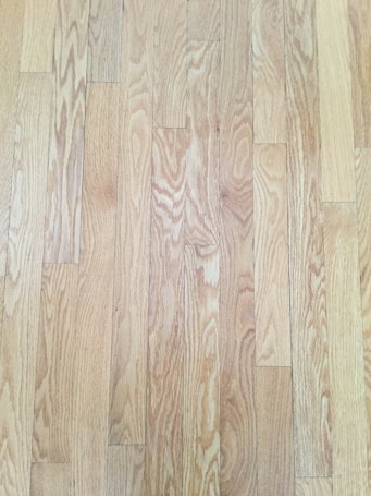 A close-up view of wooden floorboards, showing a natural wood grain pattern with varying shades of light brown and beige. The floorboards are aligned in parallel rows, exhibiting a smooth and polished surface.