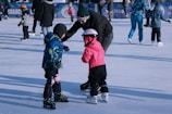 A person is helping two children who are ice skating. One child is wearing a blue and green jacket, while the other is in a pink outfit and helmet. Several other people are ice skating in the background.