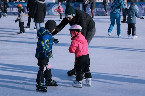 A person is helping two children who are ice skating. One child is wearing a blue and green jacket, while the other is in a pink outfit and helmet. Several other people are ice skating in the background.