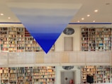 A spacious library with rows of bookshelves filled with colorful books on both levels. A striking blue and white triangular art piece is suspended from the ceiling. The room is well-lit with recessed lights, and a metal railing divides the two floors. A door and a circular painting are visible in the center.