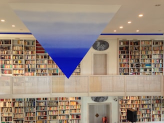 A spacious library with rows of bookshelves filled with colorful books on both levels. A striking blue and white triangular art piece is suspended from the ceiling. The room is well-lit with recessed lights, and a metal railing divides the two floors. A door and a circular painting are visible in the center.