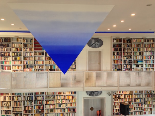 A spacious library with rows of bookshelves filled with colorful books on both levels. A striking blue and white triangular art piece is suspended from the ceiling. The room is well-lit with recessed lights, and a metal railing divides the two floors. A door and a circular painting are visible in the center.