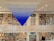 A spacious library with rows of bookshelves filled with colorful books on both levels. A striking blue and white triangular art piece is suspended from the ceiling. The room is well-lit with recessed lights, and a metal railing divides the two floors. A door and a circular painting are visible in the center.