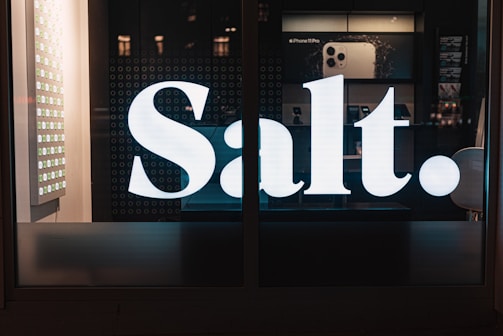 A large, illuminated 'Salt.' sign is prominently displayed on a storefront window. Behind the sign, there are displays with electronic gadgets, including images of an iPhone. The lighting is dim, creating a contrast between the glowing sign and the darker interior.