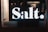 A large, illuminated 'Salt.' sign is prominently displayed on a storefront window. Behind the sign, there are displays with electronic gadgets, including images of an iPhone. The lighting is dim, creating a contrast between the glowing sign and the darker interior.