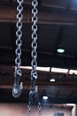 Two large industrial metal chains with hooks hang from the ceiling of a spacious warehouse. The background shows blurred structural beams and ceiling lights, indicating an industrial setting.