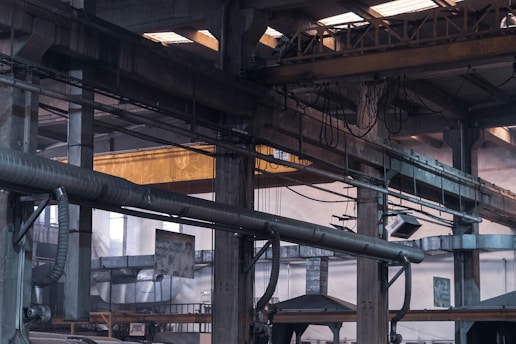 An industrial warehouse scene featuring large, metal structural beams and pipes crisscrossing the space. Overhead, a yellow crane structure with attached cables and pulleys is visible. The interior is dimly lit with natural light filtering in through skylights and windows. The image is filled with industrial textures and machinery components.