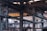 An industrial warehouse scene featuring large, metal structural beams and pipes crisscrossing the space. Overhead, a yellow crane structure with attached cables and pulleys is visible. The interior is dimly lit with natural light filtering in through skylights and windows. The image is filled with industrial textures and machinery components.