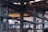An industrial warehouse scene featuring large, metal structural beams and pipes crisscrossing the space. Overhead, a yellow crane structure with attached cables and pulleys is visible. The interior is dimly lit with natural light filtering in through skylights and windows. The image is filled with industrial textures and machinery components.